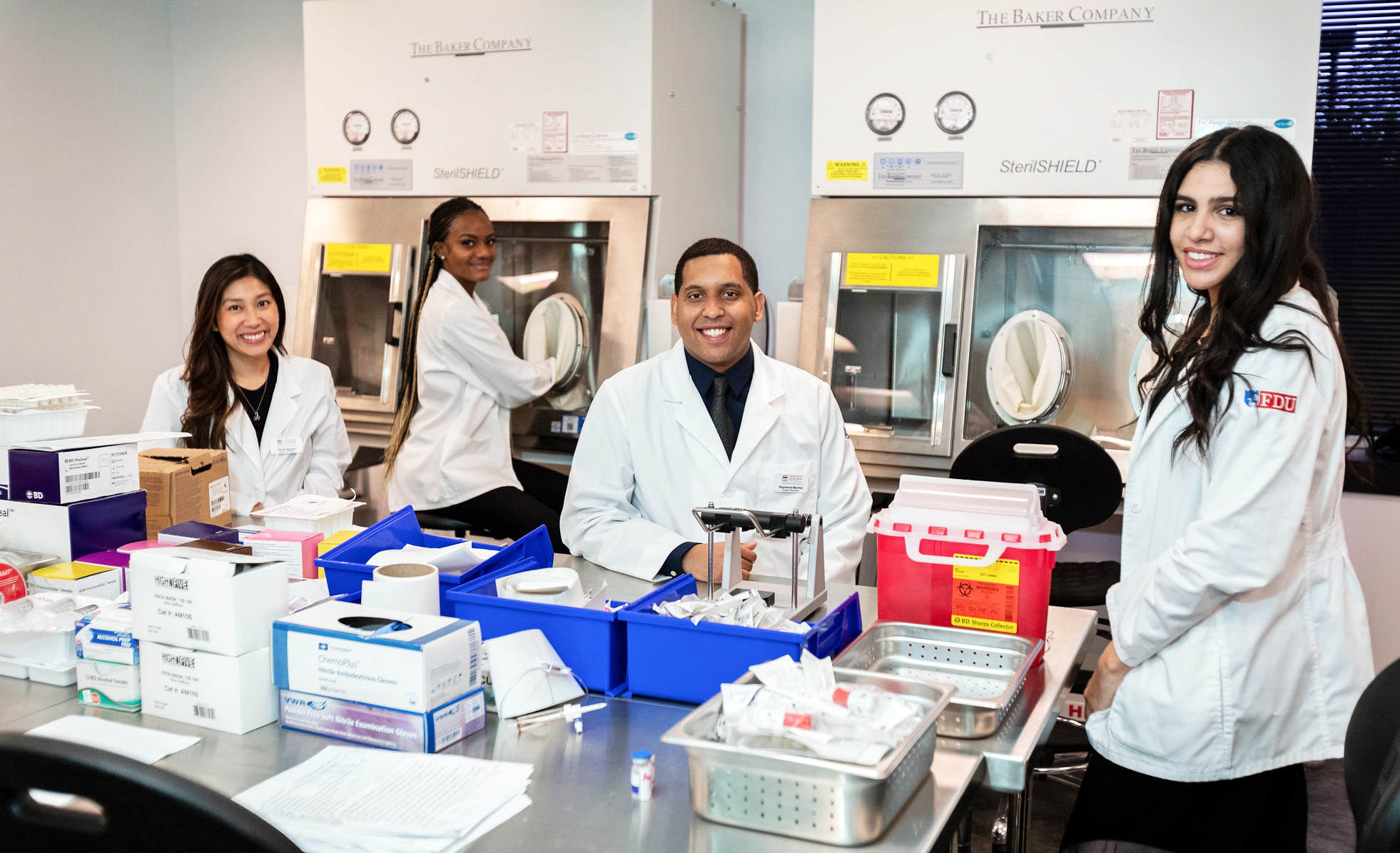 Four pharmacy students work in the Sterile Compounding lab. Pharmacy and health care equipment are on the table in front of them.