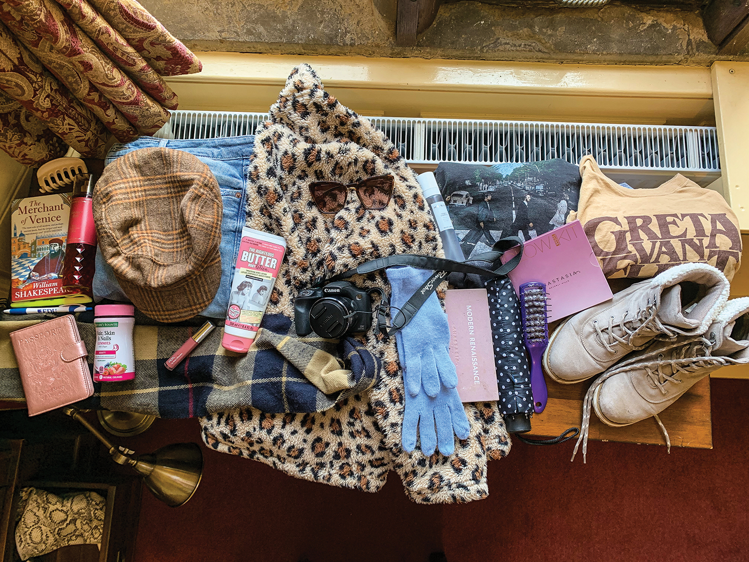 A jumble of items from a suitcase, including clothing, shoes, a camera, a book and more, sit on a wooden desk.