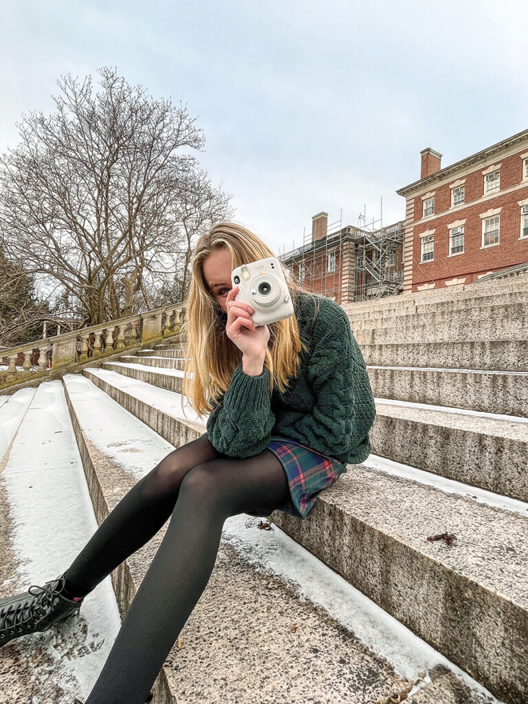 A woman sits on the back steps leading up to Hennessy Hall. She's holding up an instant camera.