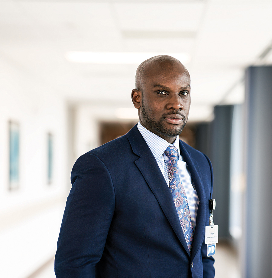 A well-dressed man in a suit and tie stands in a hospital hallway. The name on his ID badge isn't clear but it says M.D. below.