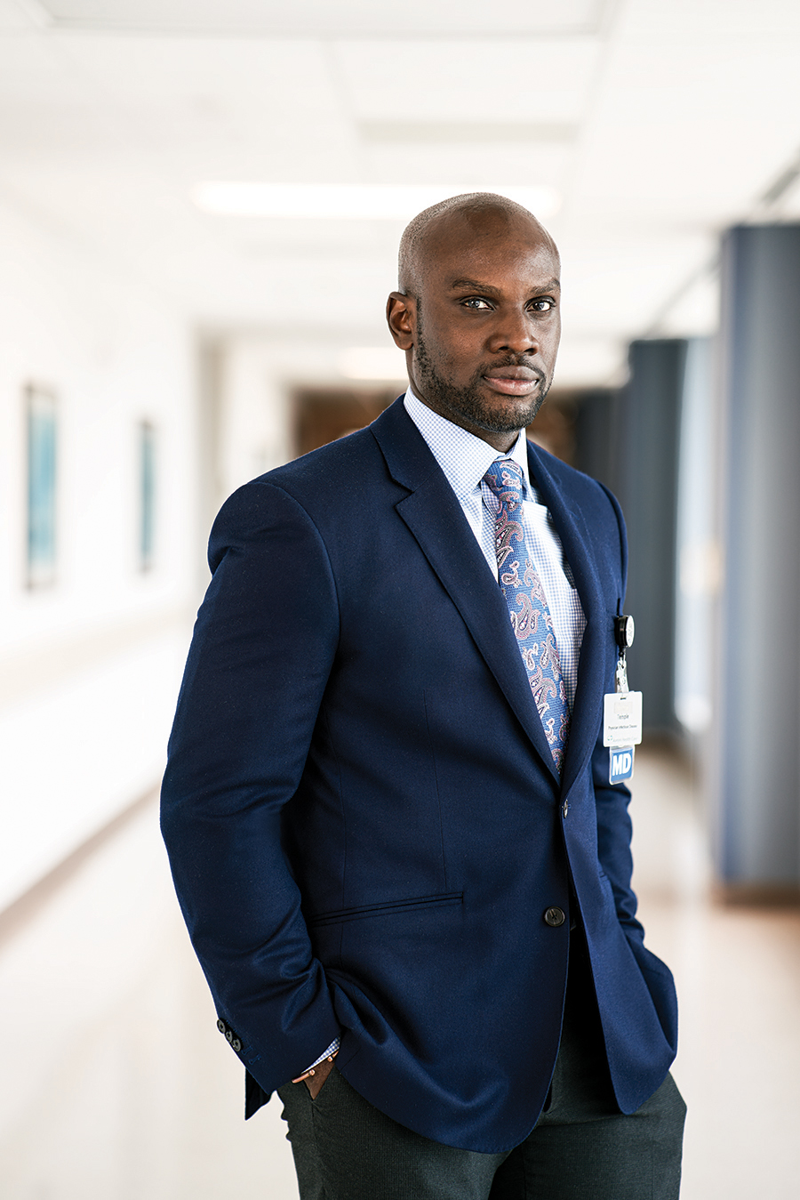 A well-dressed man in a suit and tie stands in a hospital hallway. The name on his ID badge isn't clear but it says M.D. below.