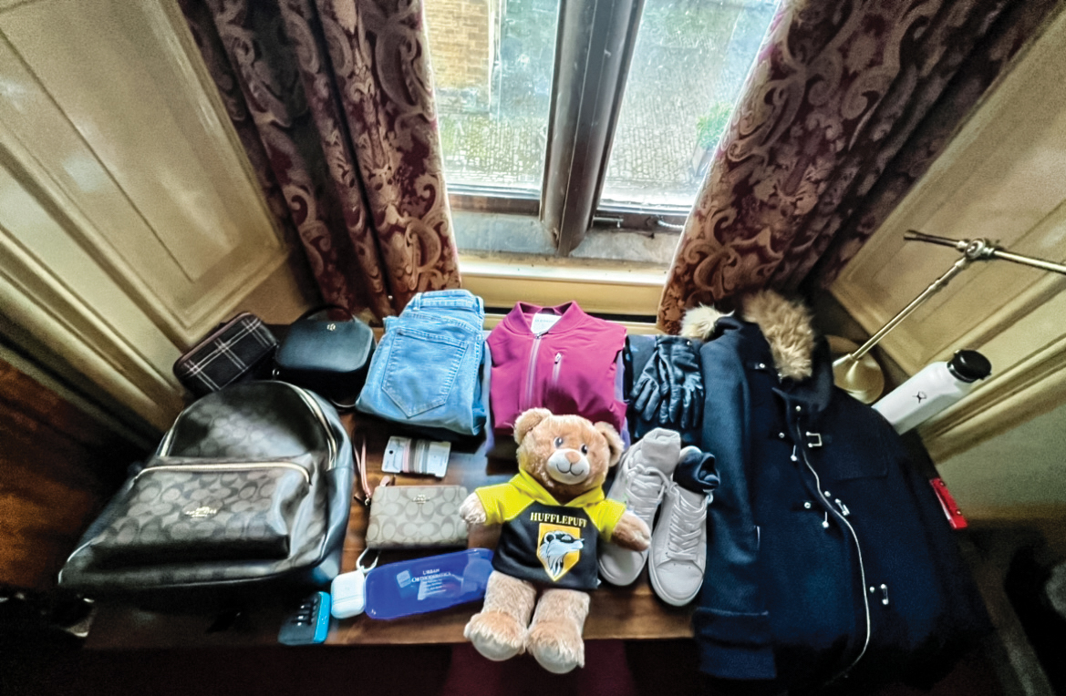 A jumble of items from a suitcase, including clothing, shoes, a teddy bear and more, arranged on a wooden desk.