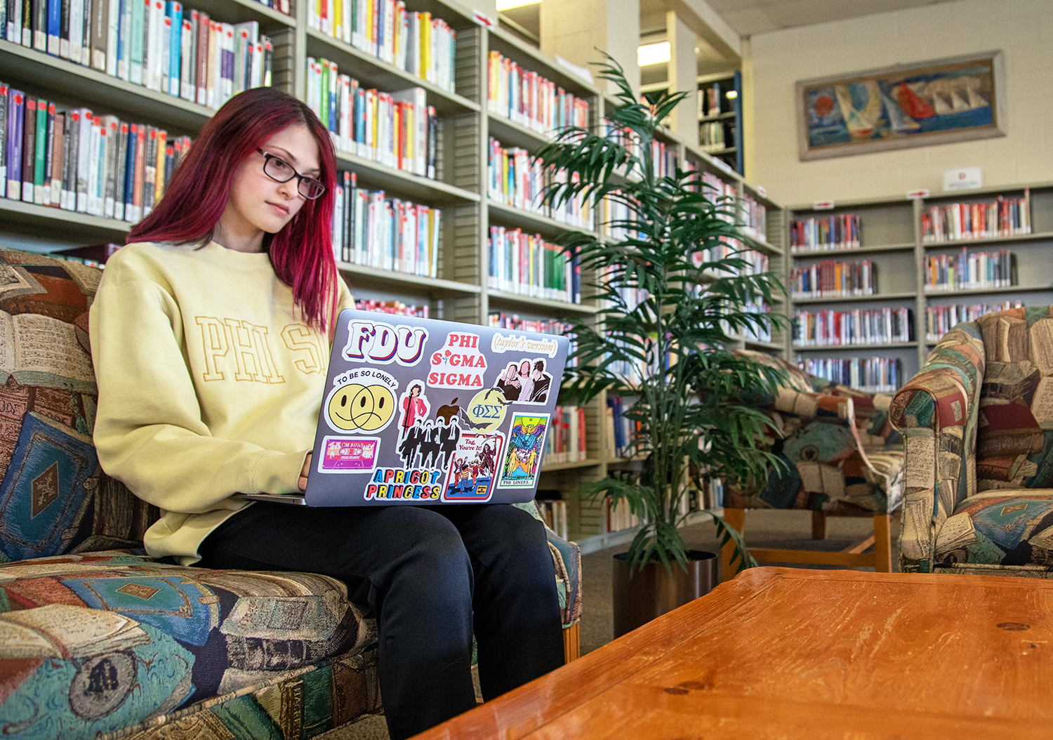 A young woman wearing glasses and sitting in the library, types on her laptop. The laptop has many colorful stickers on the exterior.