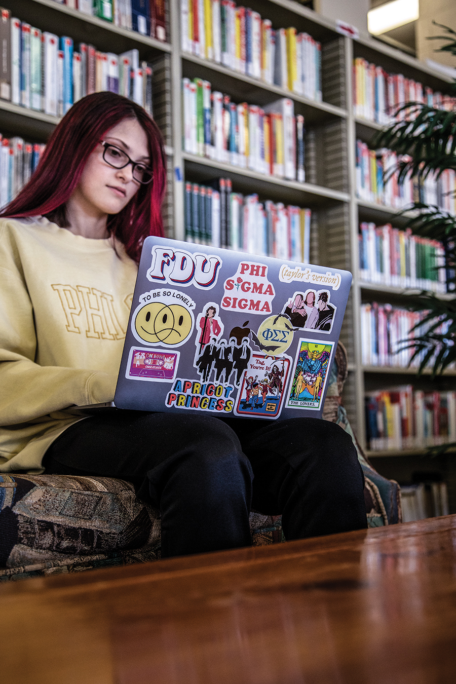 A young woman wearing glasses and sitting in the library, types on her laptop. The laptop has many colorful stickers on the exterior.
