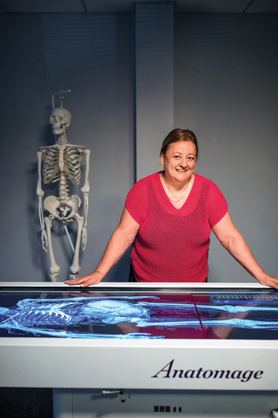 A woman leans against a table with an illuminated screen. Behind her a skeletal model hangs against the wall.