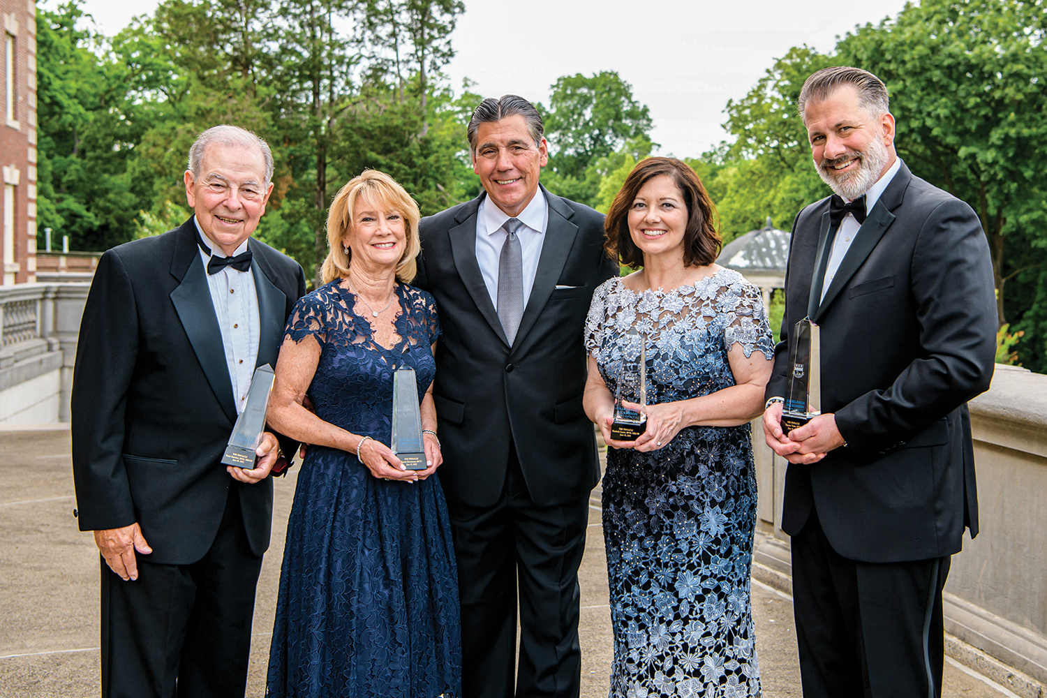 Five people, three man and two women, stand in a line, in formal wear.