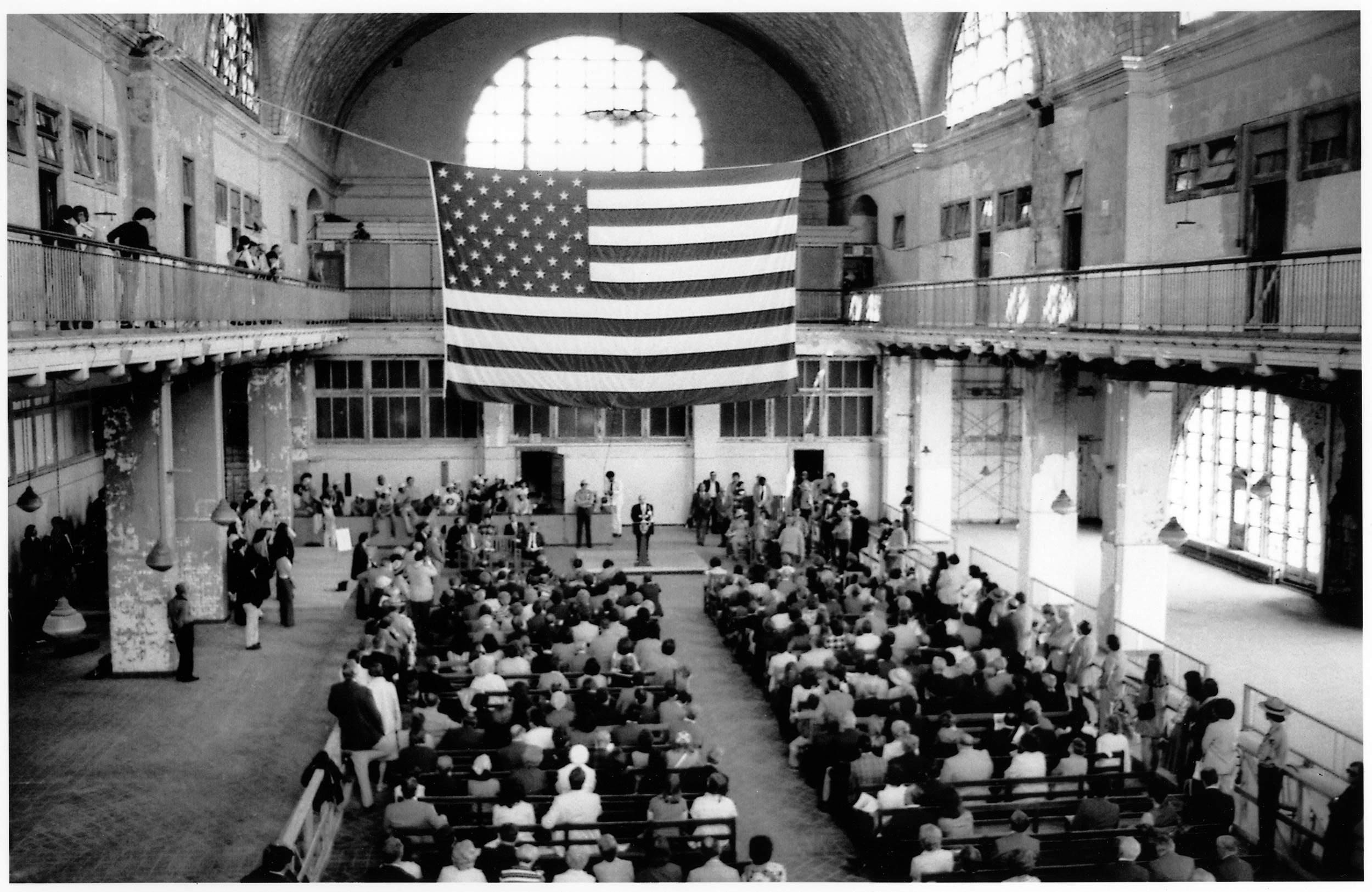 A vintage photo of a grand hall at Ellis Island, with a crowd listening to a speaker.