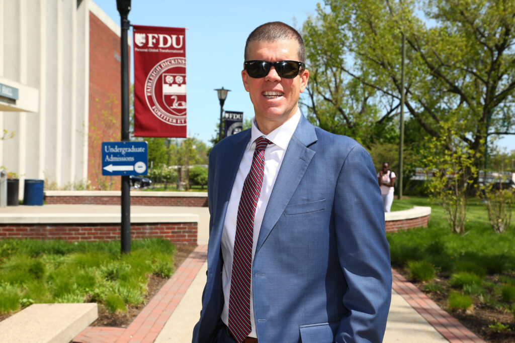 Tobin Anderson stands outside Dickinson Hall on a sunny day wearing sunglasses and a suit. 
