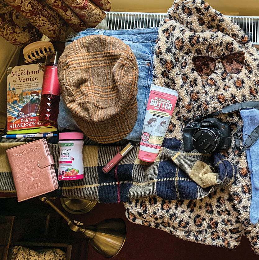 A jumble of items from a suitcase, including clothing, shoes, a camera, a book and more, sit on a wooden desk.