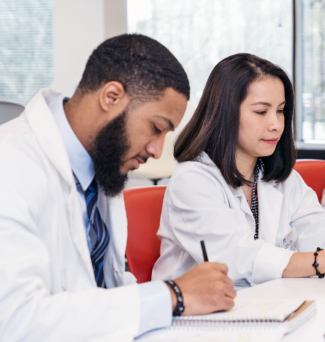 Four pharmacy students, one man and three women, all wearing their white coats, take notes and type on iPads.