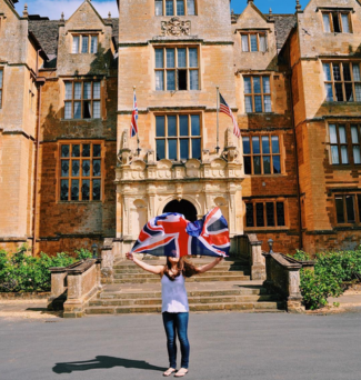a student standing in front of the Wroxton Abbey. she holds up the United Kingdom flag, covering her face.