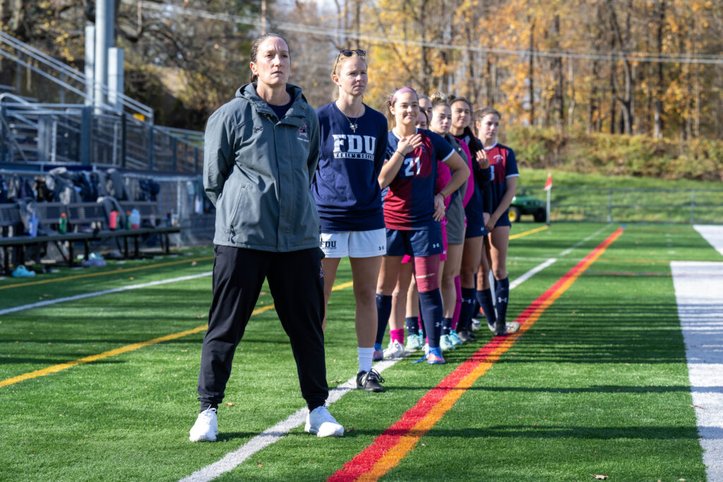 Kristin Giotta stands on a green turf field in a grey jacket and black pants with white sneakers. Standing behind her are members of the women's soccer team dressed in navy and burgundy uniforms and soccer cleats. 