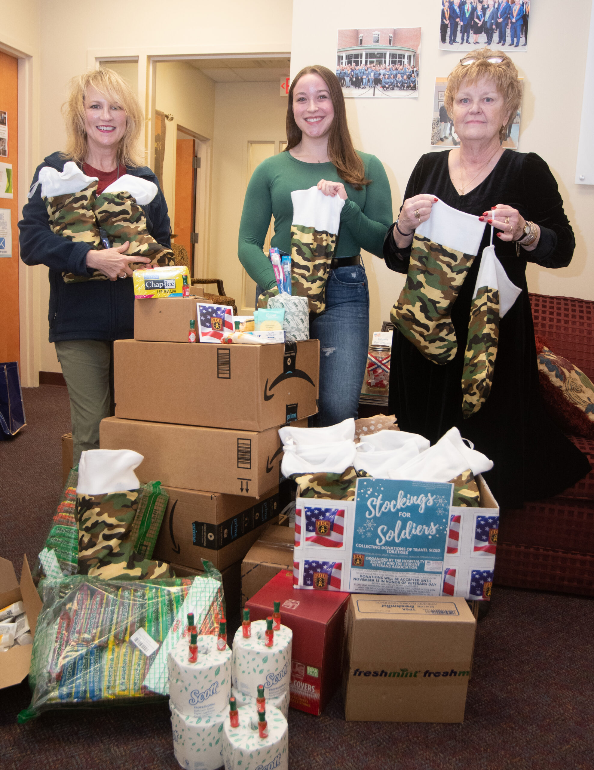 Three women stand with items donated to the Stockings for Soldiers program. 