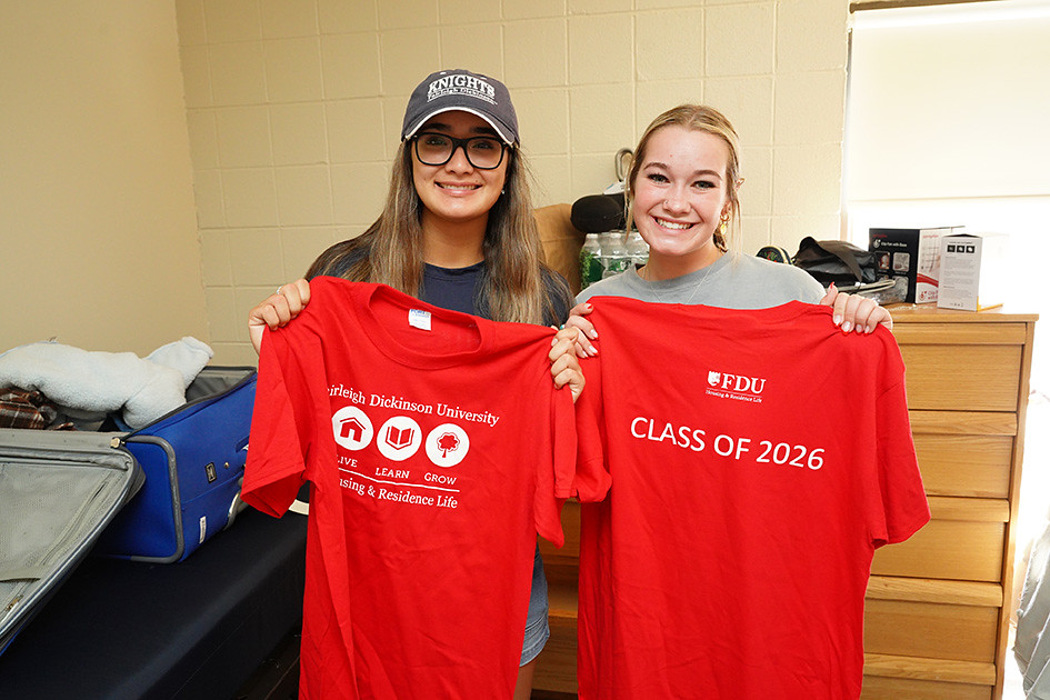 two students moving into a dorm room. they hold up shirts that read "FDU class of 2026"