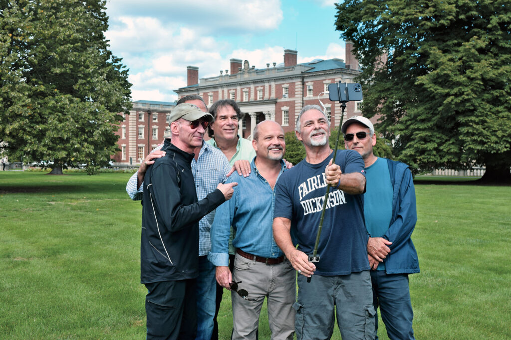 Five middle-aged men take a selfie on the lawn in front of Hennessy Hall.