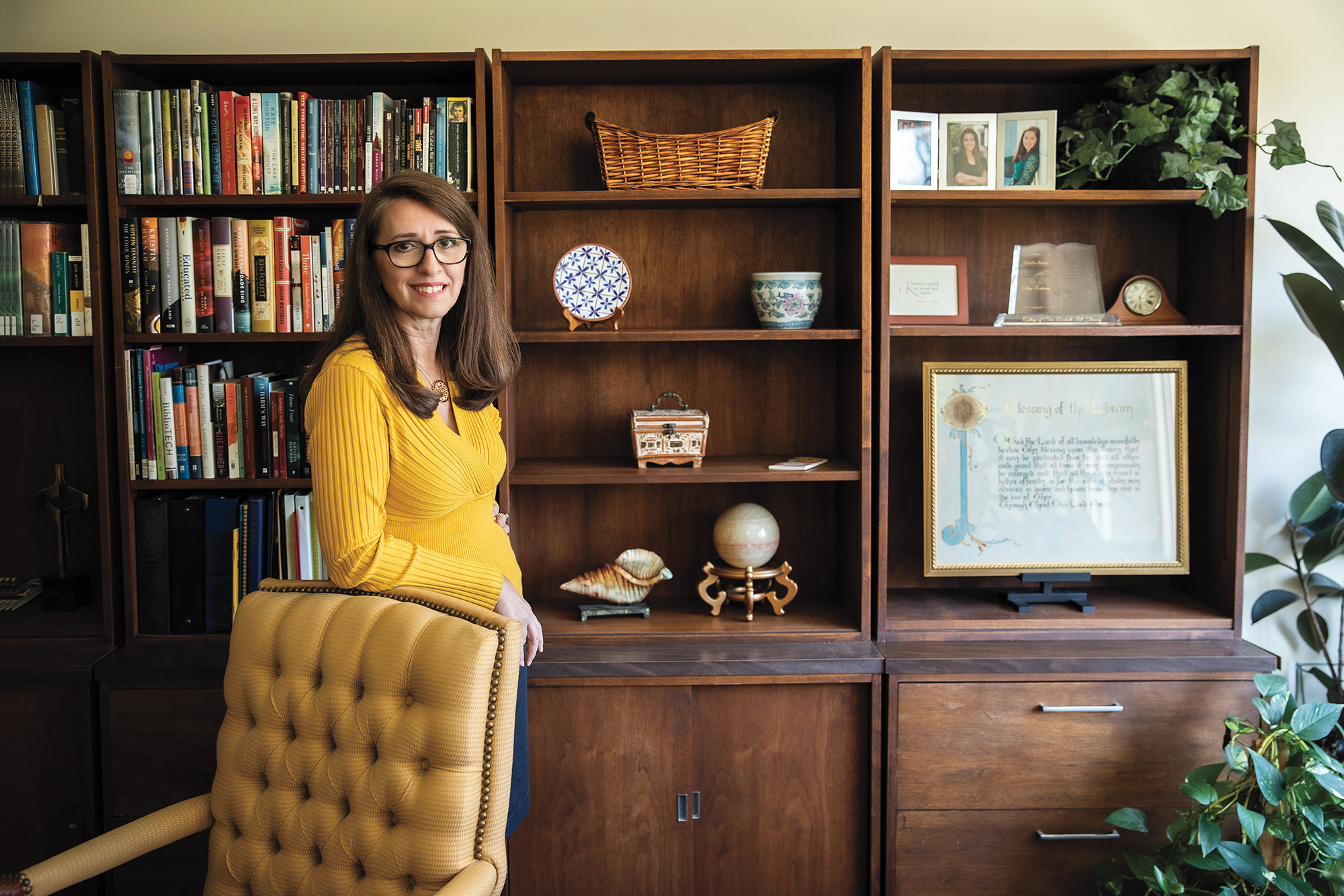 A woman wearing glasses rests her arm on the top of a chair. Behind her, bookcases filled with books, ceramics and framed art.