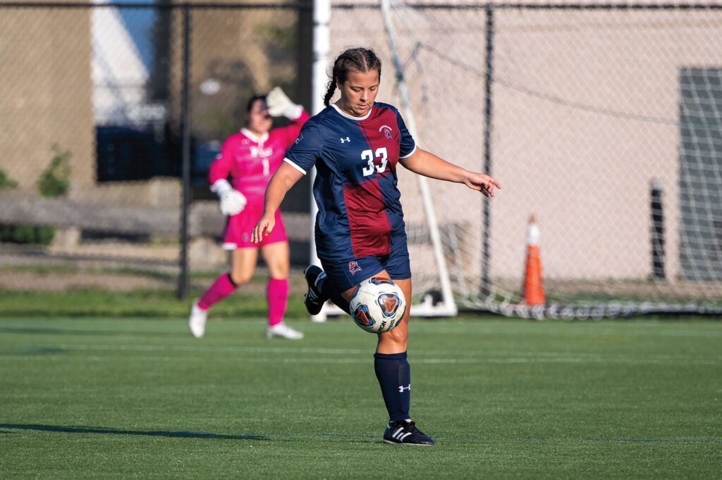 A female soccer player in a burgundy and blue uniform prepares to kick a soccer ball on a green turf field. 