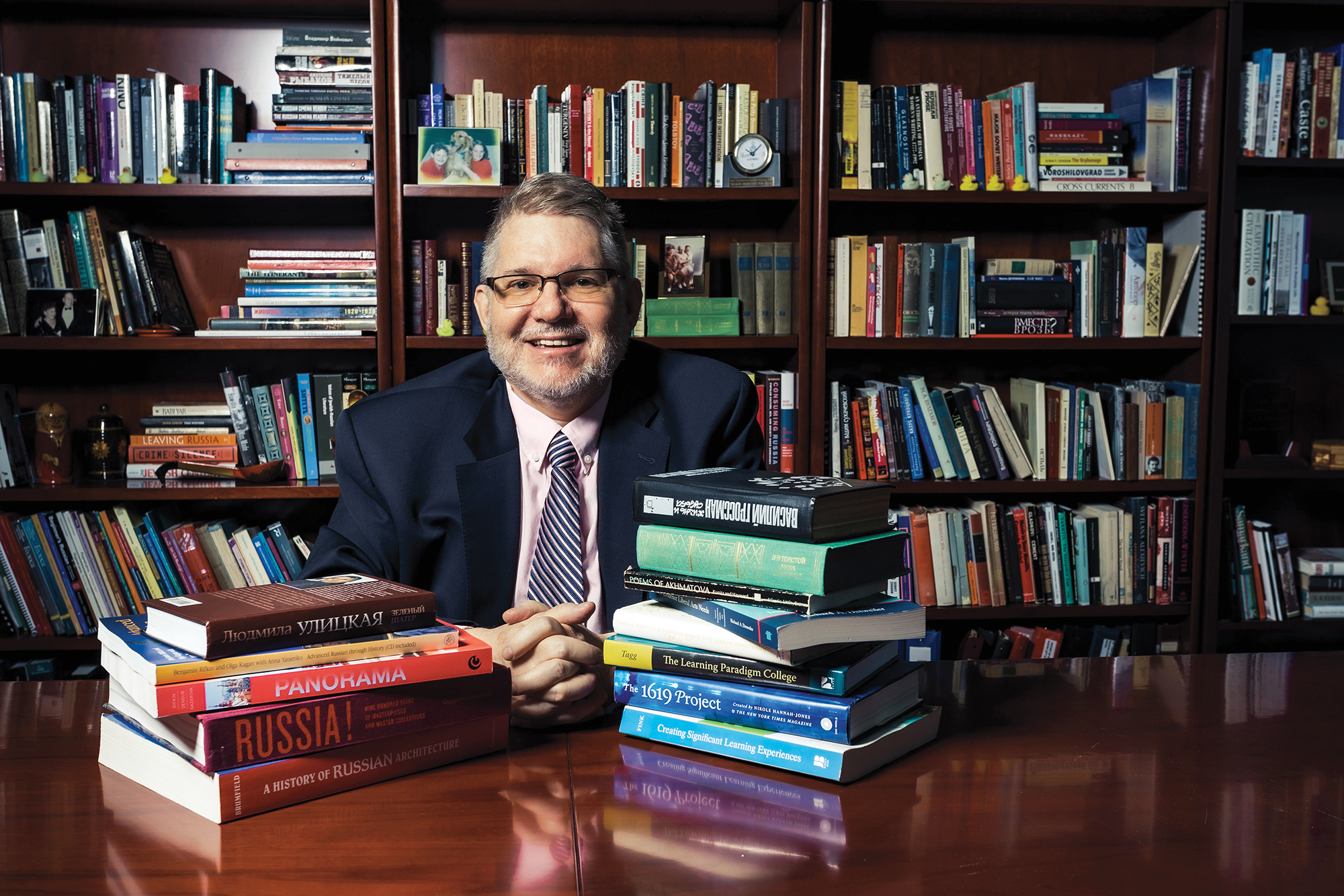 A man wearing glasses sits at a table with stacks of books on each side of him. Behind the man, a bookcase full of books spans the wall. 