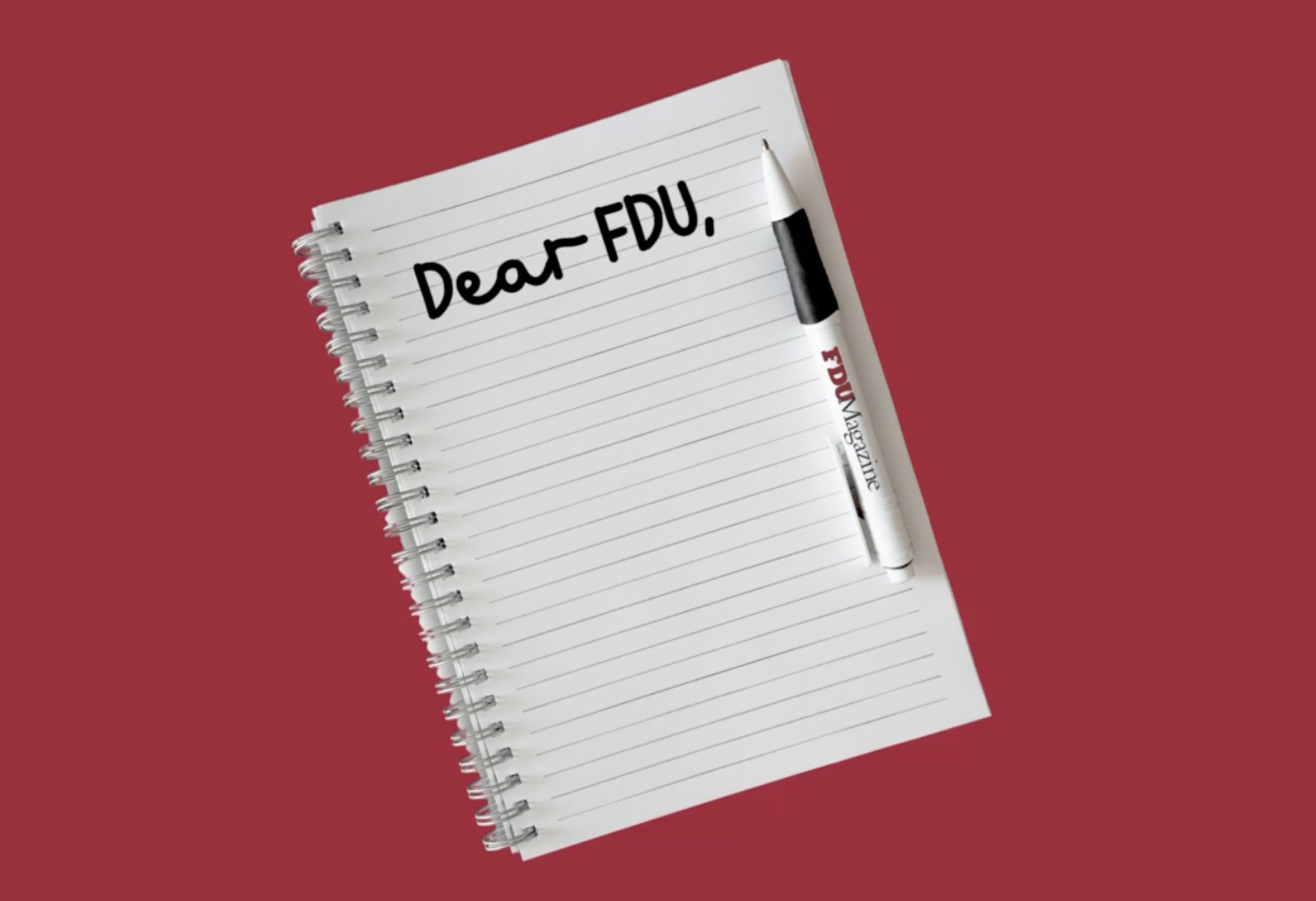 A red background with a spiralbound notebook with an FDU-branded pen. At the top of the page, handwriting says, 