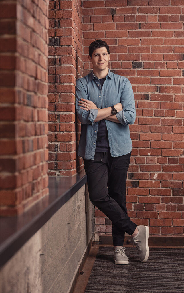 A young man casually leans against an interior brick wall, arms and legs crossed.