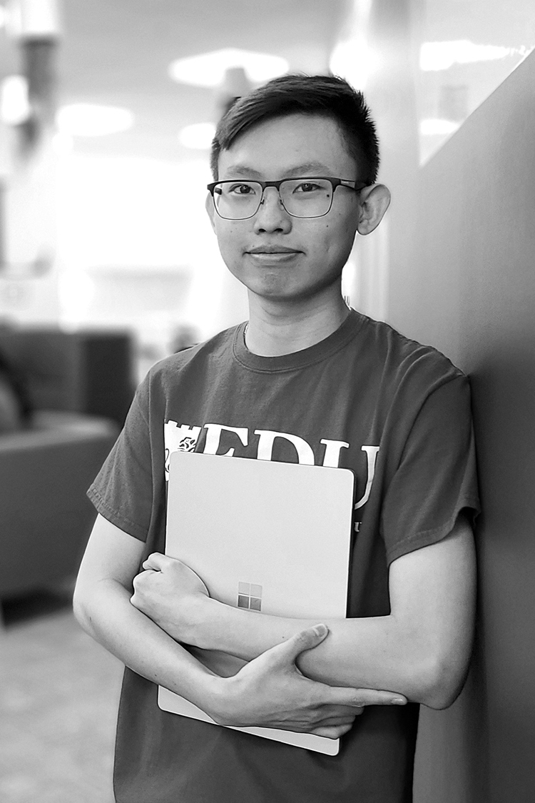 A young man wearing glasses and holding a folder, leans against a wall.