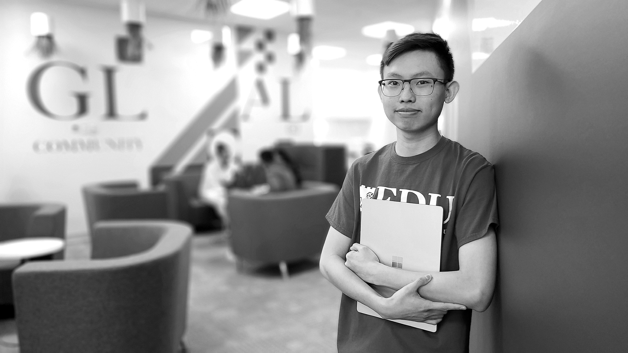 A young man wearing glasses and holding a folder, leans against a wall.