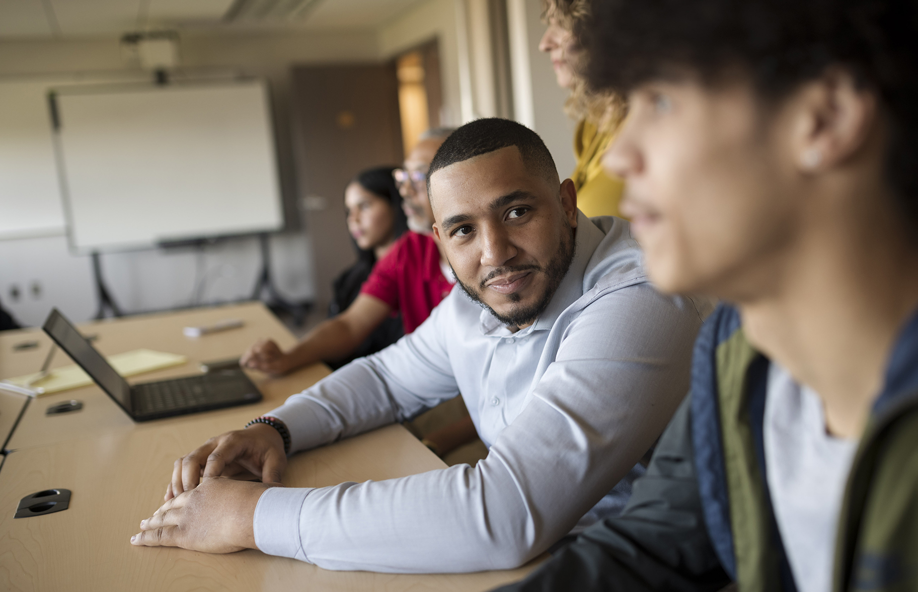 A male student turns his attention to a peer. The two are sitting at a table.