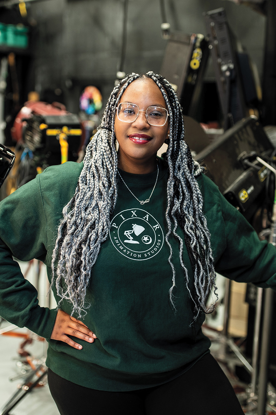 A young woman wearing a Pixar sweatshirt stands among lights and other film equipment on set.