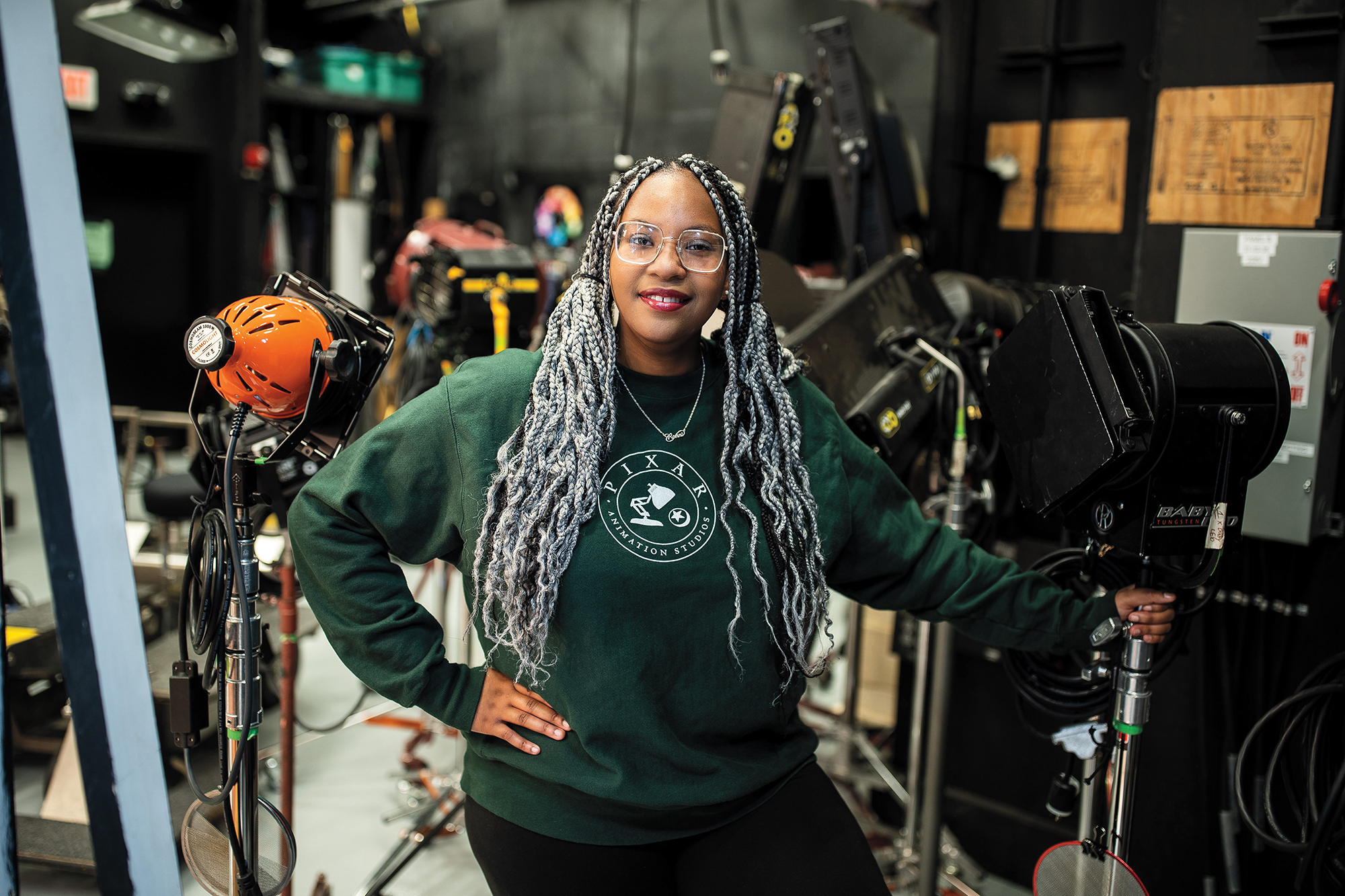 A young woman wearing a Pixar sweatshirt stands among lights and other film equipment on set.