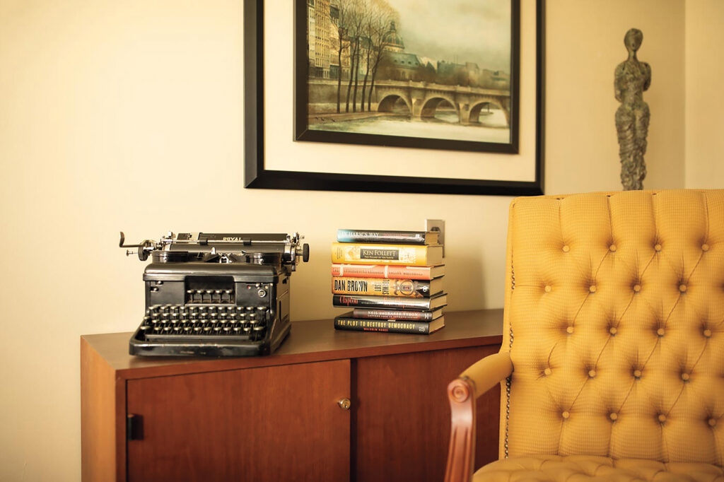 A sideboard with an old-fashioned typewriter and book stack. An armchair is next to the sideboard.