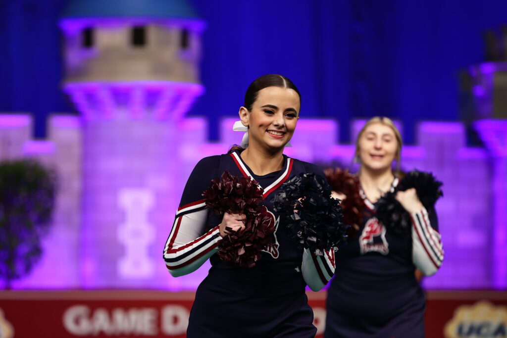 A female cheerleader smiles as she claps her pom poms together. 