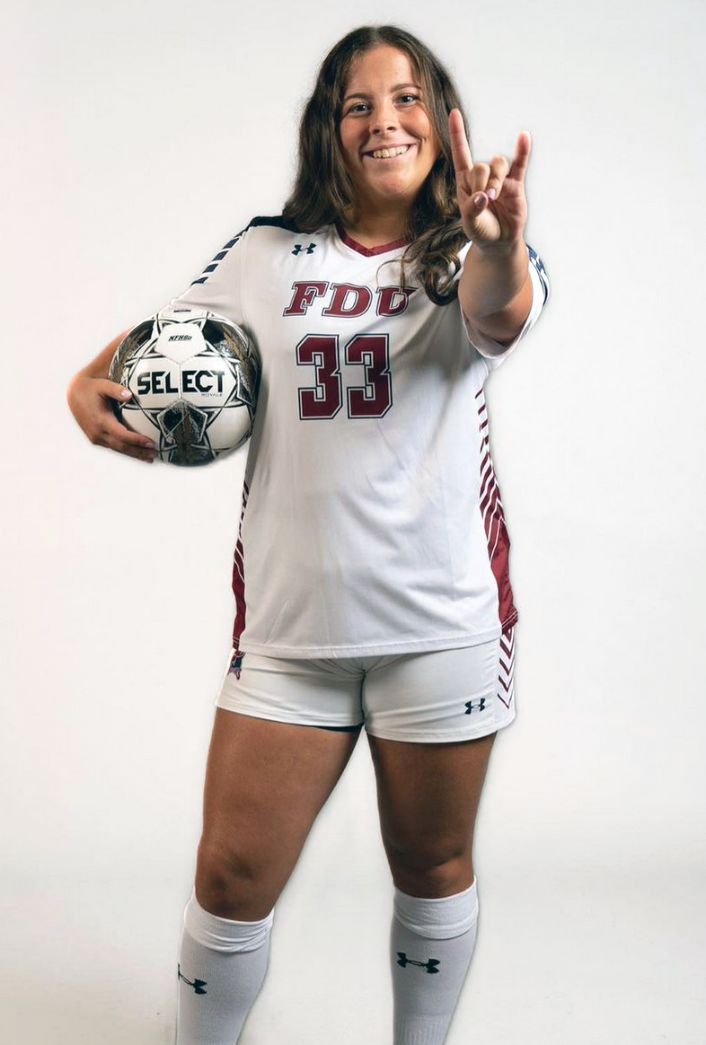 A young woman stands holding a soccer ball under one arm and makes devil horns with her other hand.