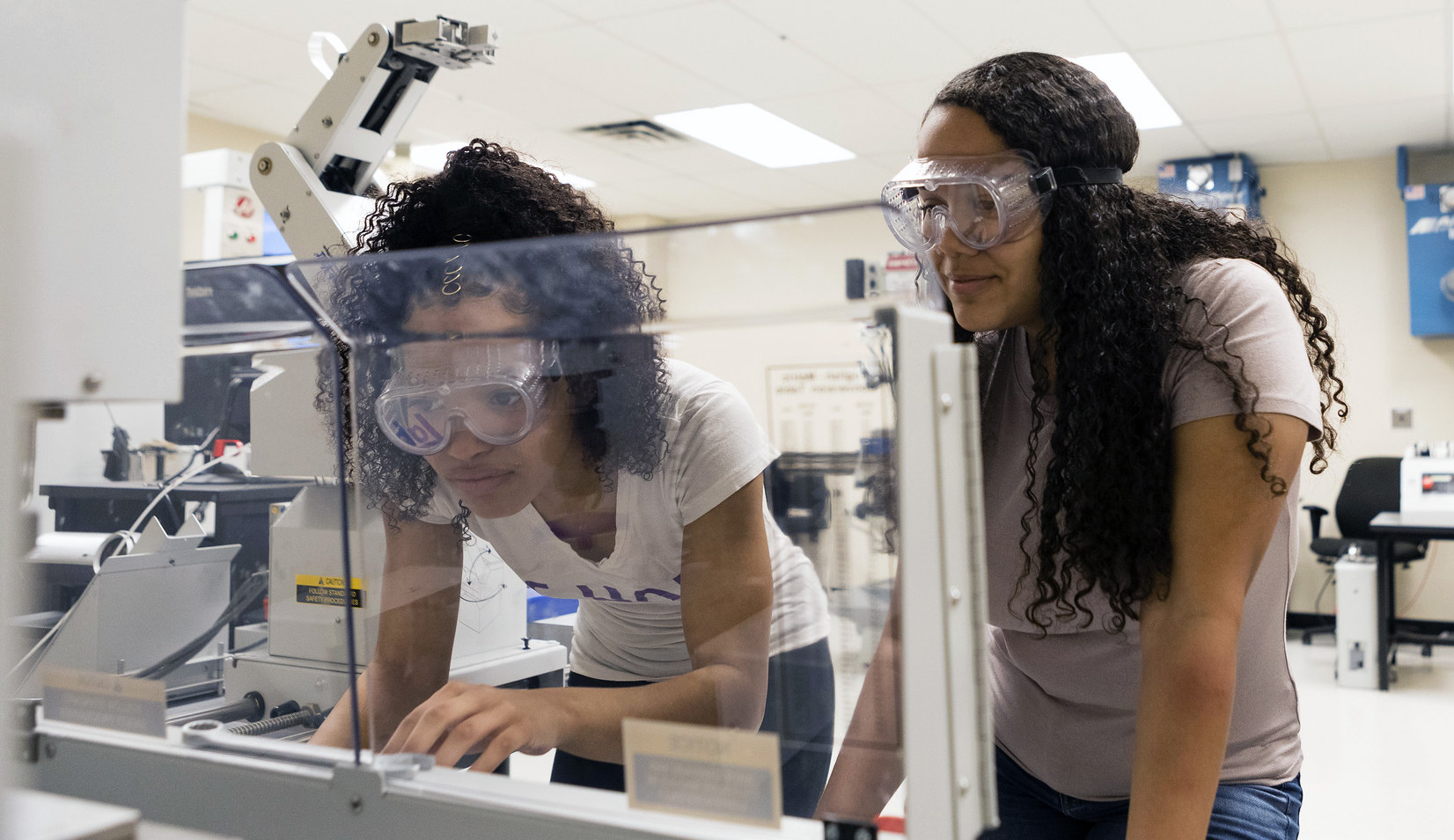 Two female engineering students wear googles and investigate something in the lab.
