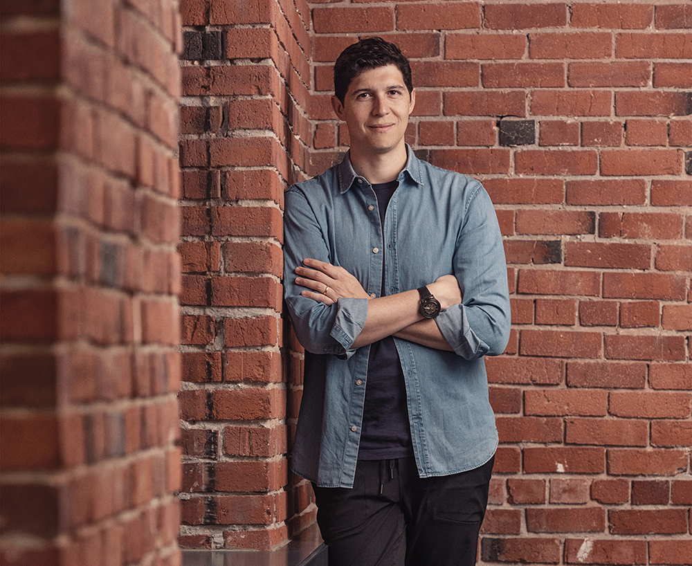 A young man casually leans against an interior brick wall, arms and legs crossed.