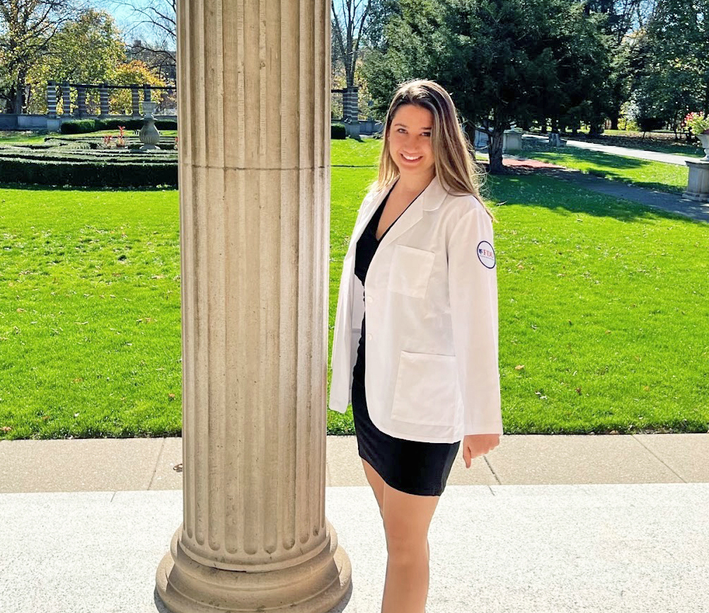 A young woman in a white coat poses for a photo on the portico of Hennessy Hall.