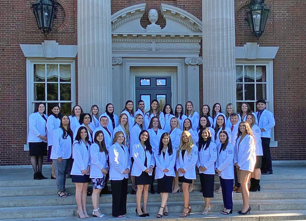 The first cohort of physician assistant studies students poses on the steps of Hennessy Hall in their white coats.