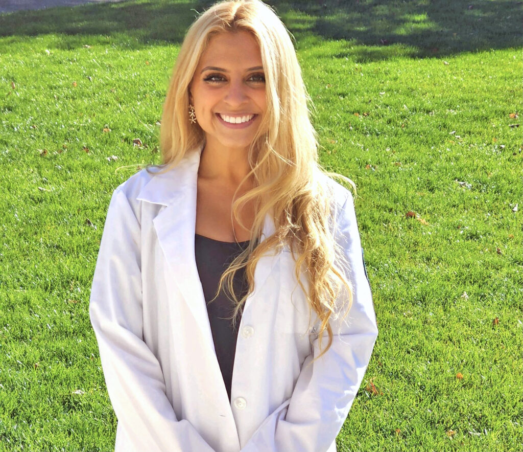 A young woman wearing a white coat stands outside and smiles.