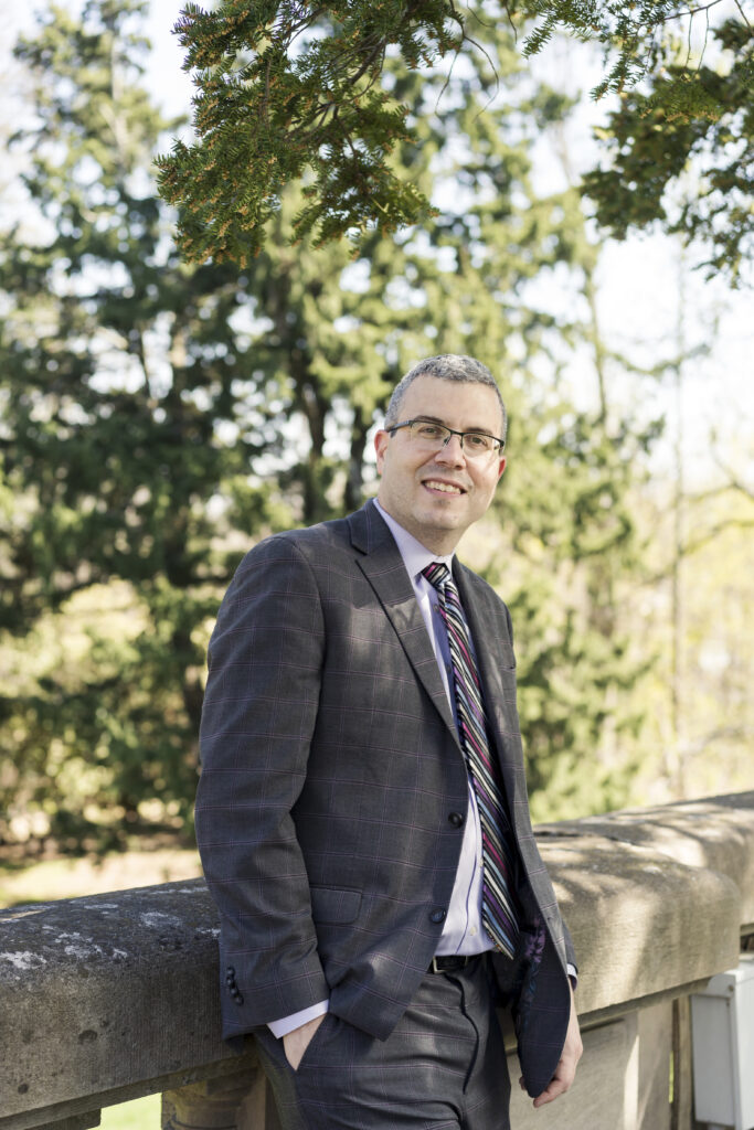 FDU President Michael J. Avaltroni stands outside against a stone wall in a grey suit wearing a purple shirt and tie