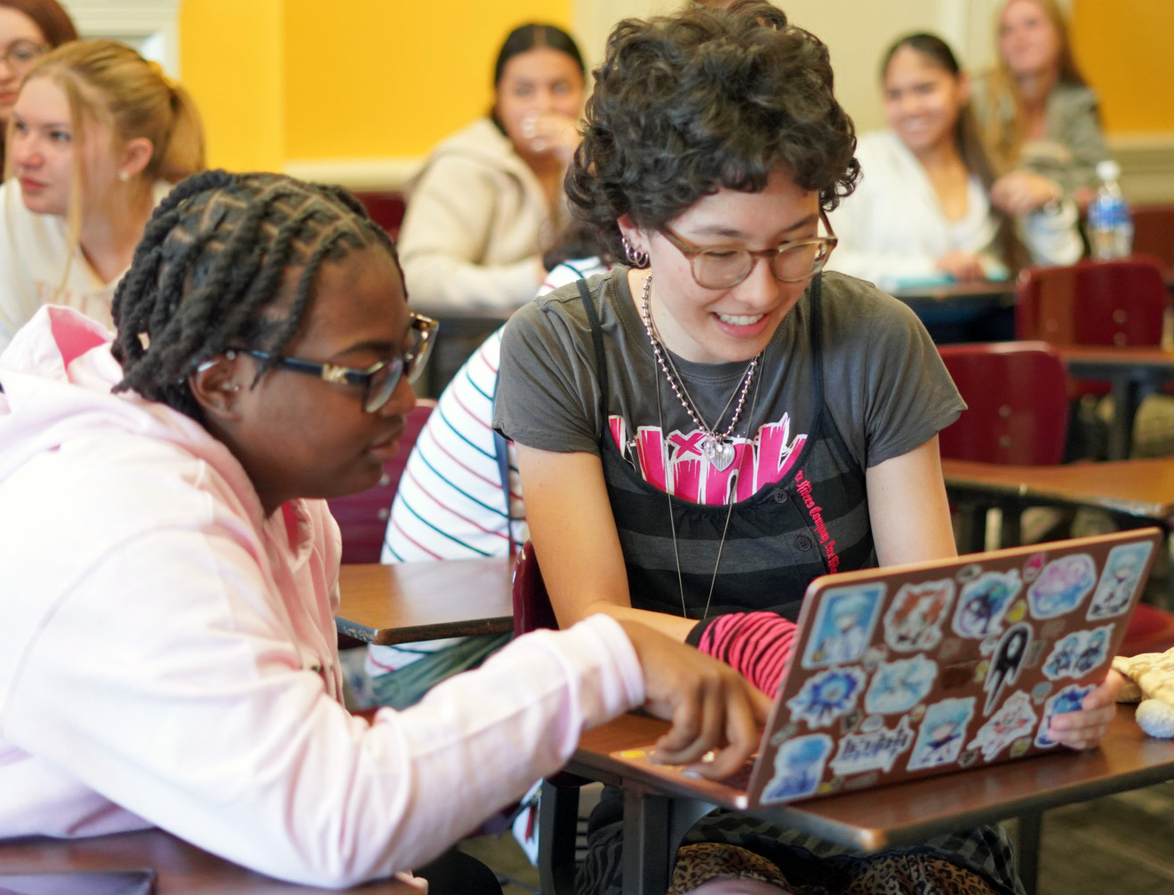 Two young women, both wearing glasses, lean over to look at a laptop screen. 