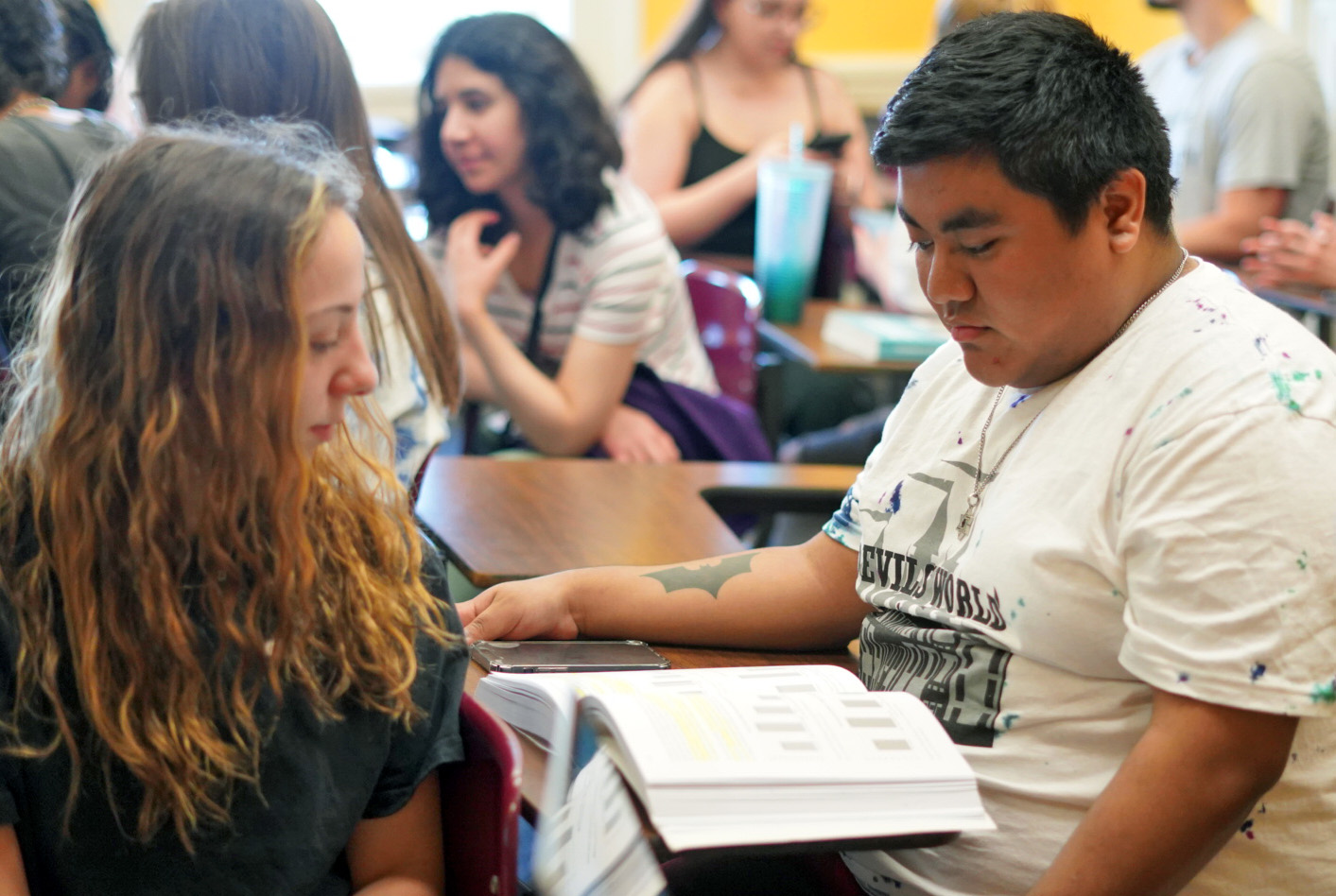 A male student sits at a desk, with a textbook open to face him. A female student turns in her seat to look behind her at the book.