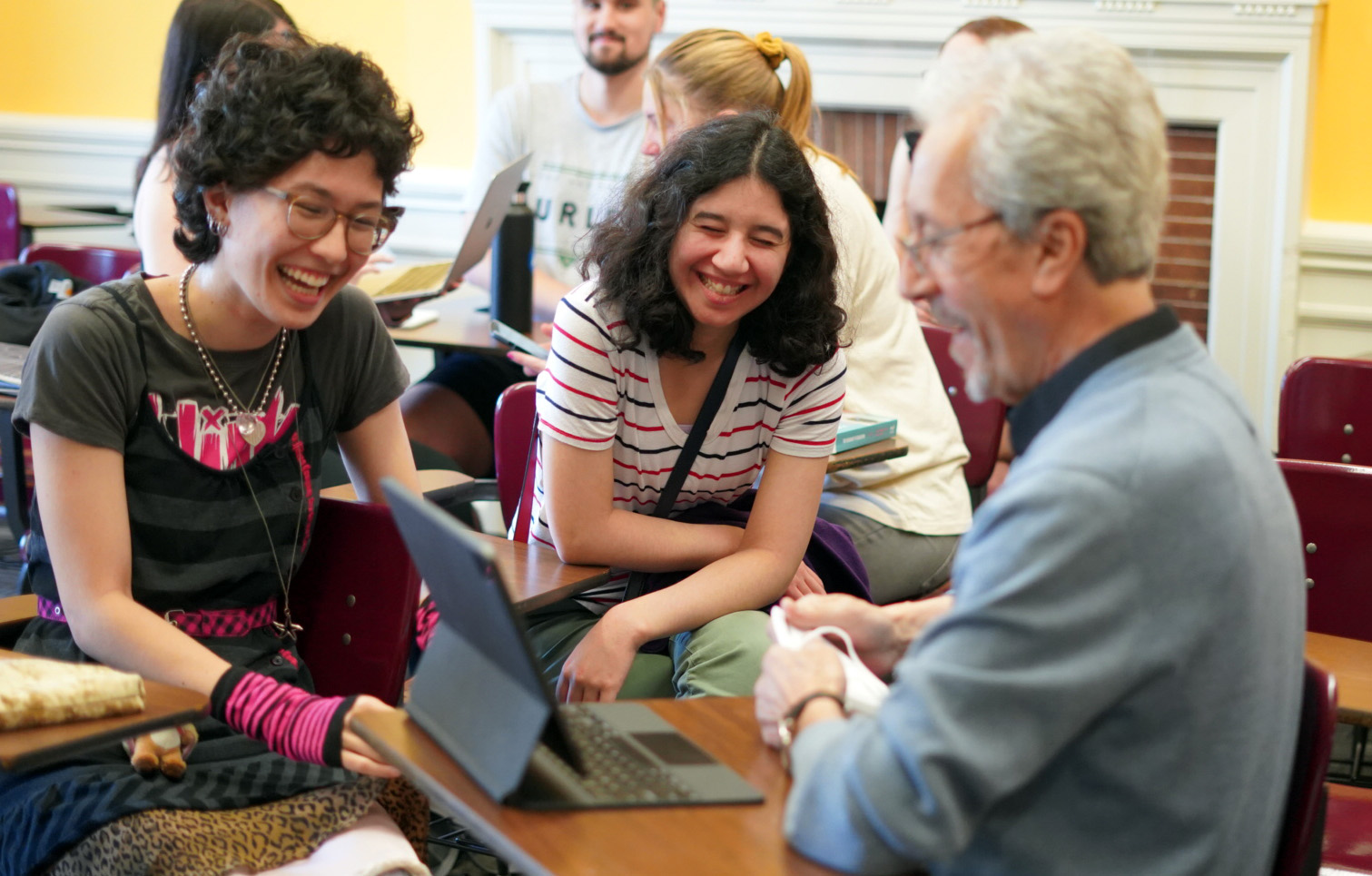 Two female students laugh while in conversation with a male professor. All three sit at desks in a classroom.