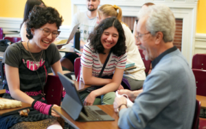 Two female students laugh while in conversation with a male professor. All three sit at desks in a classroom.