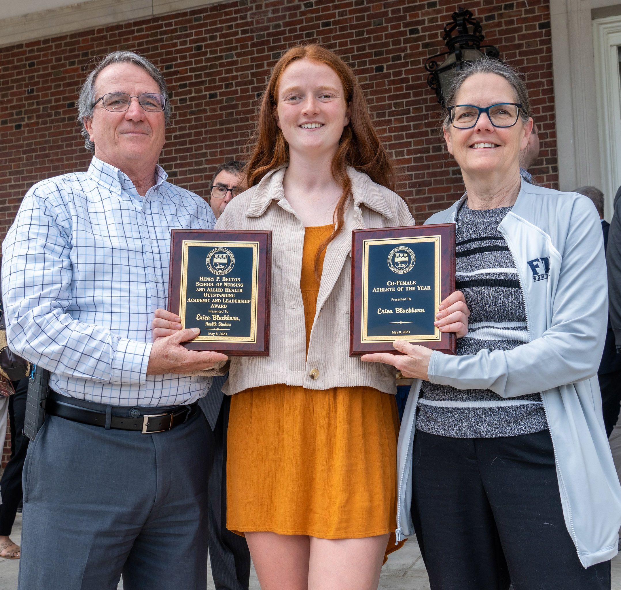 A young woman, flanked by a man and a woman, holds up an award plaque.