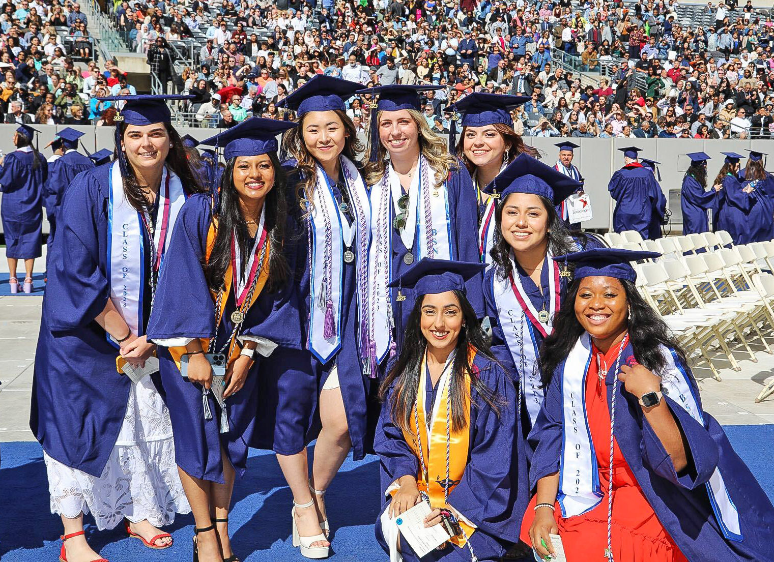A group of female graduations, wearing caps, gowns and regalia, on the field at Commencement.