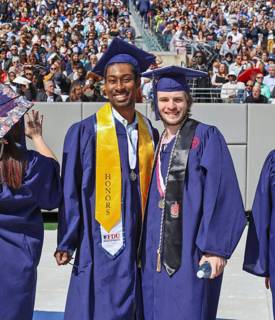 Two male graduates wearing graduation regalia smile.