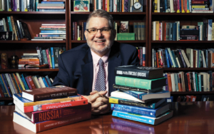 A man wearing glasses sits at a table with stacks of books on each side of him. Behind the man, a bookcase full of books spans the wall.