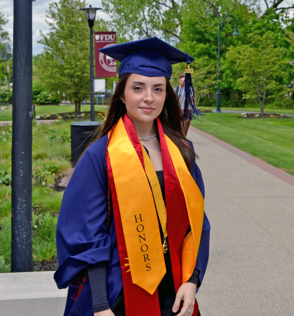 A young woman wears academic regalia — a cap, gown, cords and sashes — and stands outside.