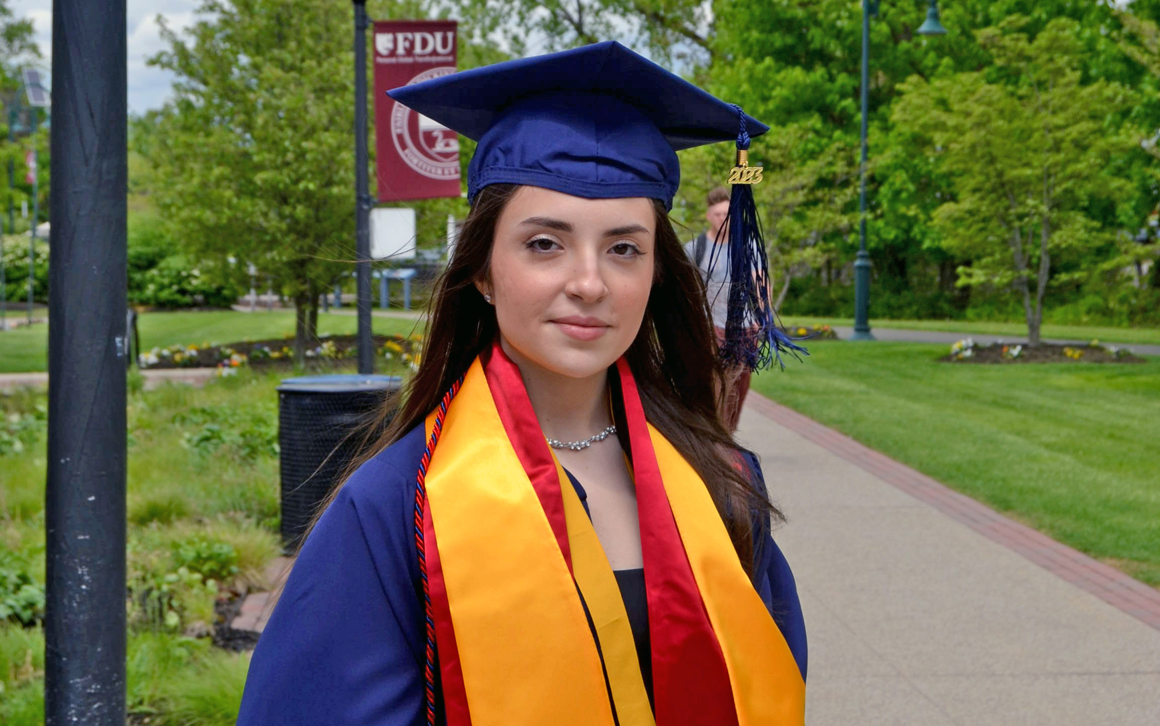 A young woman wears academic regalia — a cap, gown, cords and sashes — and stands outside.