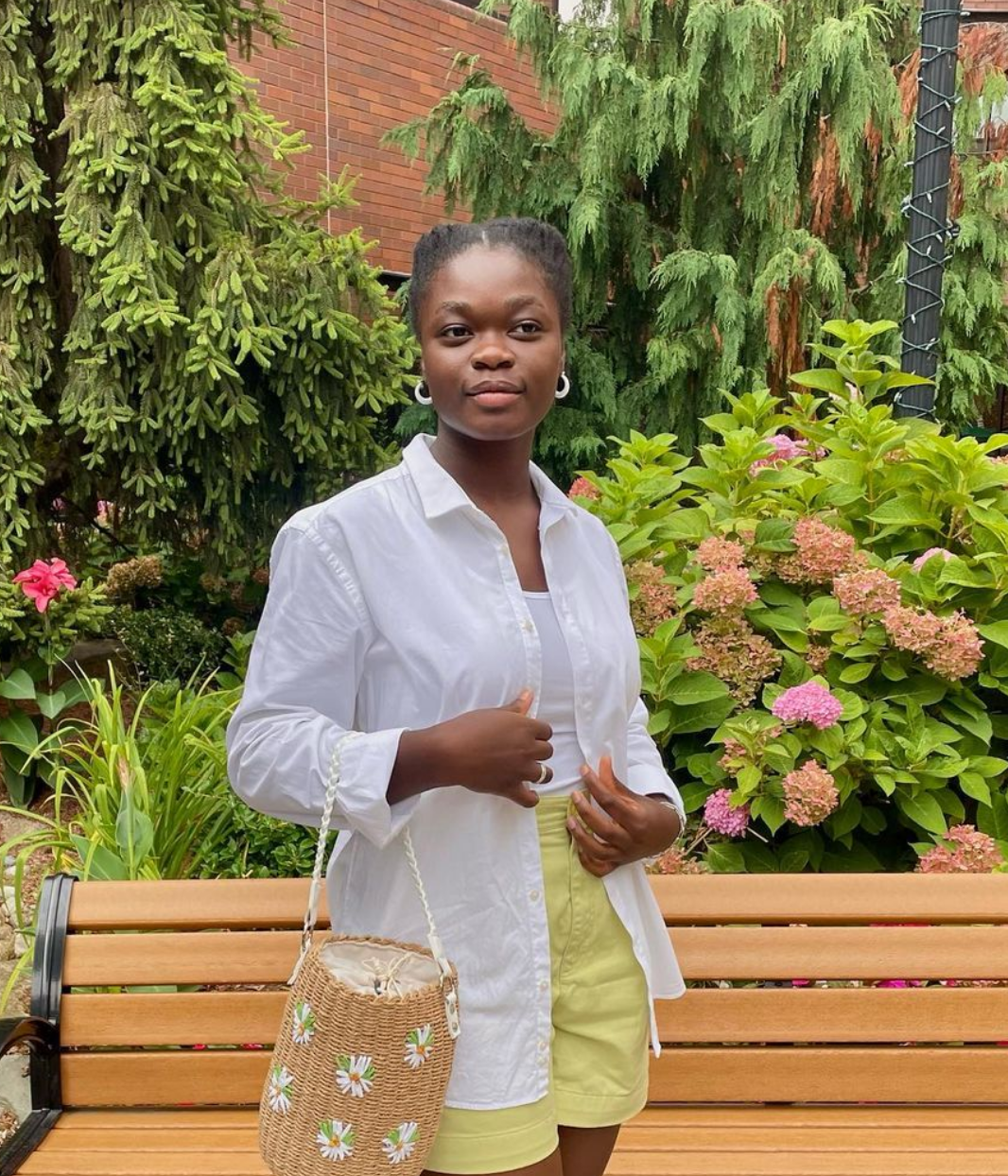 A young woman stands outside, near a garden, in front of bench and holds a purse.