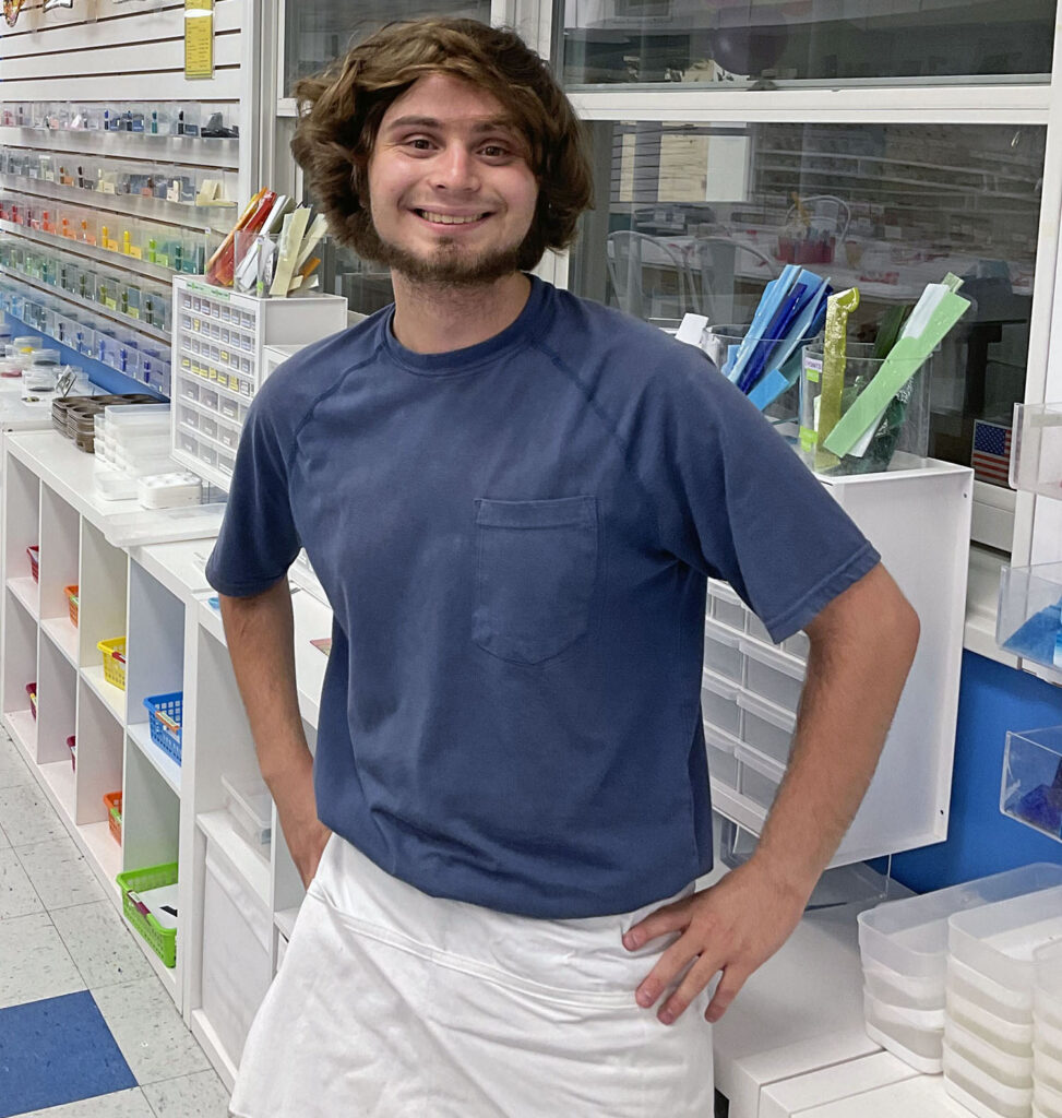 A young man wearing an art apron poses for a photo in front of mosaic glass.